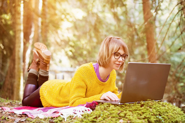 Toned image of a freelancer girl portrait in a yellow sweater and glasses looking thoughtfully at the laptop screen in the nature in a coniferous forest. The concept of freelancing freedom for travel - Photo, Image