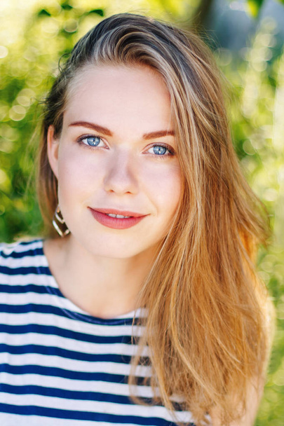 mooie lachende witte Kaukasische meisje vrouw met lang blond haar en blauwe ogen dragen gestreept t-shirt buiten in de zomer park tussen groen gebladerte bomen in de camera kijken. - Foto, afbeelding