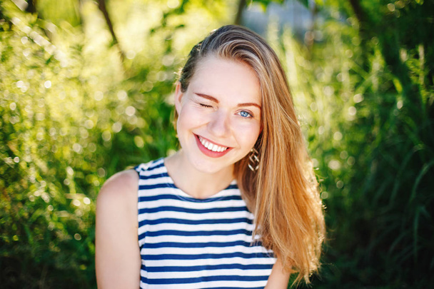 mooie glimlachend ergens wit Kaukasische meisje vrouw met lang blond haar en blauwe ogen dragen gestreept t-shirt buiten in de zomer park tussen groen gebladerte bomen - Foto, afbeelding