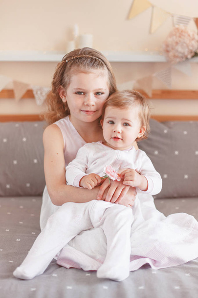 Portrait of cute adorable white Caucasian girl holding little sister baby on laps knees. Smiling children sitting together on bed in bedroom looking in camera. Sibling love - Fotoğraf, Görsel