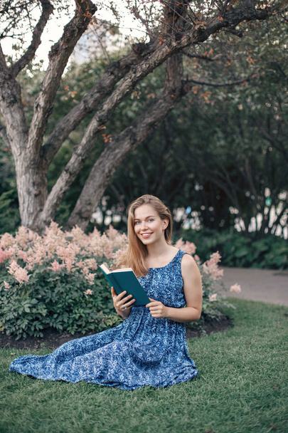 Beautiful smiling white Caucasian girl woman with long blonde hair in long dress sitting on grass in park outside reading book. Lifestyle hobby concept - Photo, image