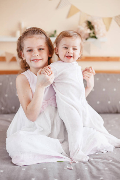 Portrait of cute adorable white Caucasian girl holding little sister baby on laps knees. Smiling children sitting together on bed in bedroom looking in camera. Sibling love - Foto, Imagem