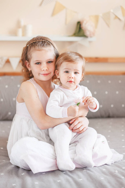 Portrait of cute adorable white Caucasian girl holding little sister baby on laps knees. Smiling children sitting together on bed in bedroom looking in camera. Sibling love - Photo, Image