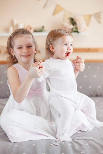 Portrait of cute adorable white Caucasian girl holding little sister baby on laps knees. Smiling children sitting together on bed in bedroom looking in camera. Sibling love - Foto, Imagem