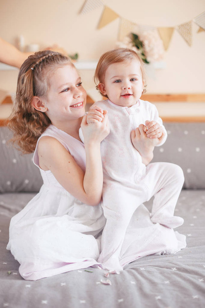 Retrato de linda adorable chica blanca caucásica sosteniendo bebé hermana pequeña en las rodillas vueltas. Niños sonrientes sentados juntos en la cama en el dormitorio mirando a cámara. Amor hermano
 - Foto, imagen