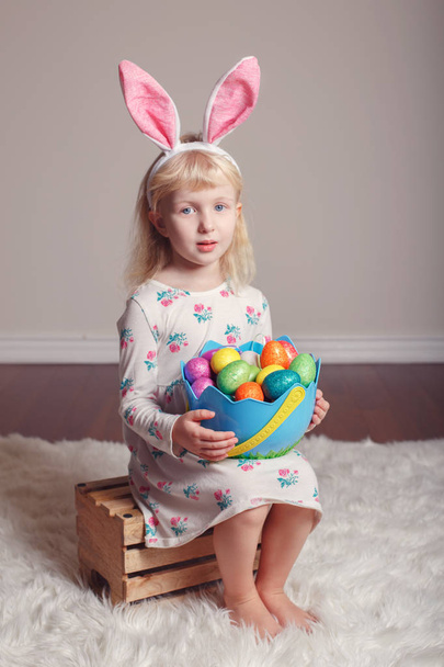 Милая очаровательная белая девочка с кроличьими ушами пасхального кролика, сидящая на деревянной коробке в студии. Ребенок держит праздничную корзину с красочными яйцами, празднуя традиционный христианский праздник
  - Фото, изображение