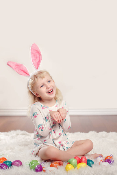Cute adorable funny Caucasian child girl wearing Easter bunny rabbit ears sitting on soft fluffy rug carpet in studio. Kid with holiday colorful eggs playing celebrating traditional Christian holiday  - Photo, Image