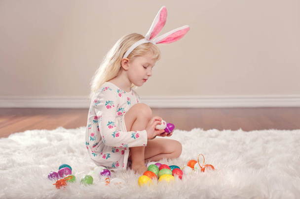 Cute adorable Caucasian child girl wearing Easter bunny rabbit ears sitting on soft fluffy rug carpet in studio. Kid holding holiday colorful eggs celebrating traditional holy Christian holiday  - Photo, Image