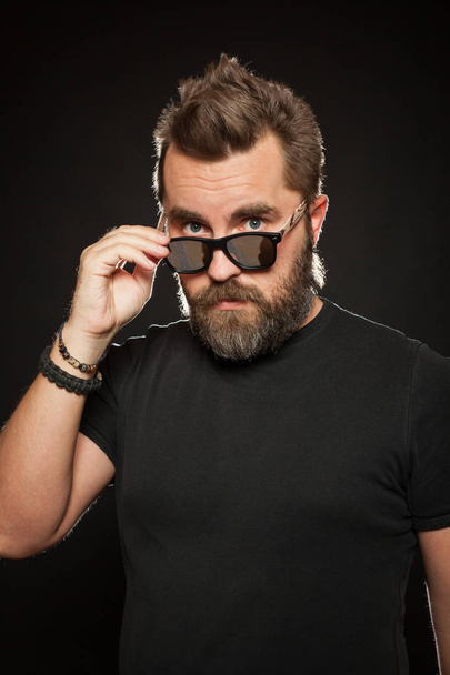 A handsome, strong man with a stylish hairstyle and beard dresses sunglasses in the Studio on a black background - Foto, immagini