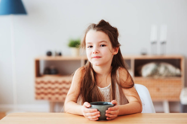 happy 5 years old kid girl having breakfast at home in the morning. Sitting at table with cup with tea or hot cocoa, healthy life concept in modern scandinavian interior - Foto, immagini