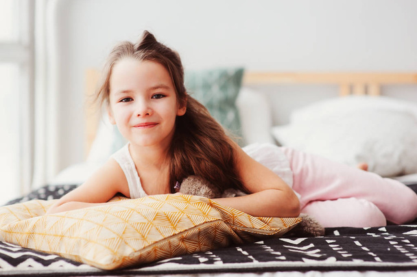 happy little child girl lying on her bed in the morning, waking up in comfortable room with modern bedlinen and pillows, cozy homely scene  - Фото, изображение