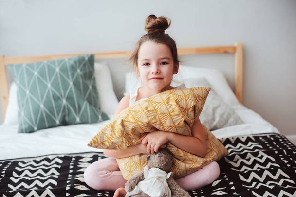 happy child girl sitting on bed and hugs pillow, waking up in early morning or going to sleep. Kid in comfortable room enjoying weekend - Valokuva, kuva