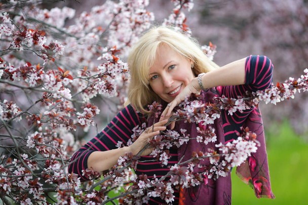 Portrait of happy smiling Caucasian blond woman with long hair near blossoming plum cherry tree, no teeth, looking to the camera - Photo, Image