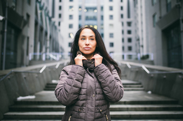 portrait of a woman on a business theme. Young caucasian brunette with long hair girl in long jacket, coat stands on business center background, office building with glass facade. Overcast in winter - Фото, изображение