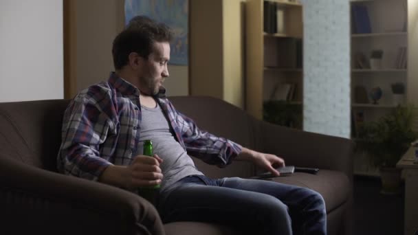 Guy sitting on sofa with beer bottle in hand, using remote control to switch TV - Imágenes, Vídeo