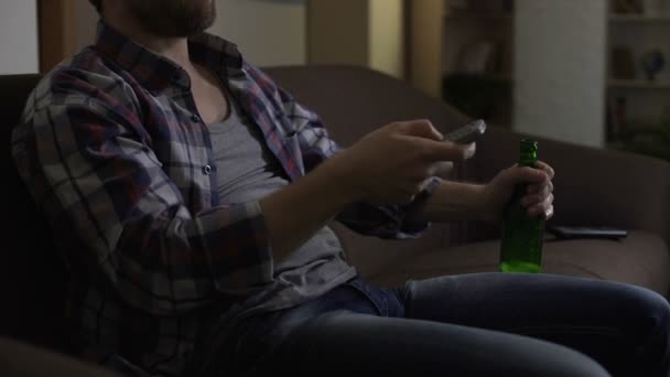 Man holding bottle of beer on knee, switching TV channels with remote control - Felvétel, videó