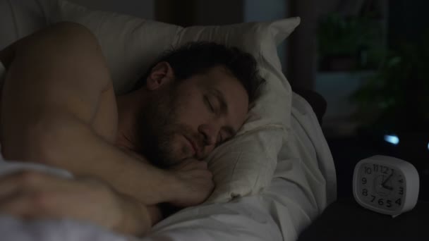 Bearded man sleeping on sofa bed, clock standing on night table, late night rest - Filmati, video
