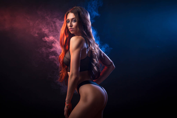 Young dark-haired woman in black lingerie posing against a background of red and   blue smoke from a vape on a black isolated background - Fotó, kép