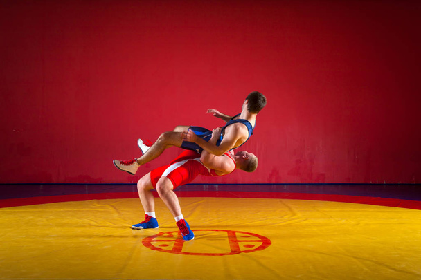 Two strong wrestlers in blue and red wrestling tights are wrestlng and making a  hip throw wrestling on a yellow wrestling carpet in the gym. Young man doing grapple. - Photo, Image