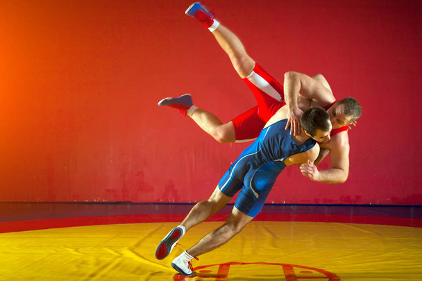 Zwei starke Wrestler in blauen und roten Wrestling-Strumpfhosen ringen und werfen sich in der Turnhalle auf einen gelben Wrestling-Teppich. Junger Mann macht Rangelei. - Foto, Bild