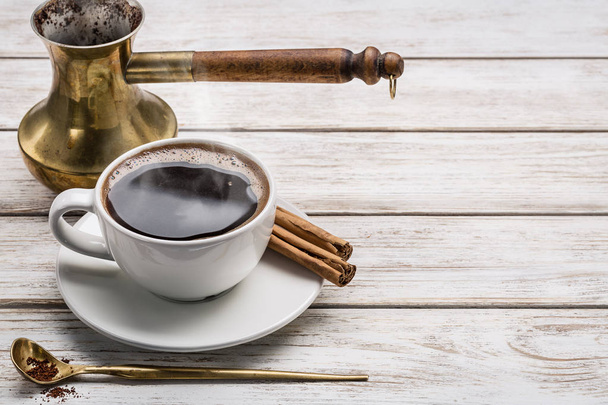 White cup of coffee and cinnamon tubes, spoon with ground coffee, coffee pot (turka) on white wooden table. Copy space. - Photo, image