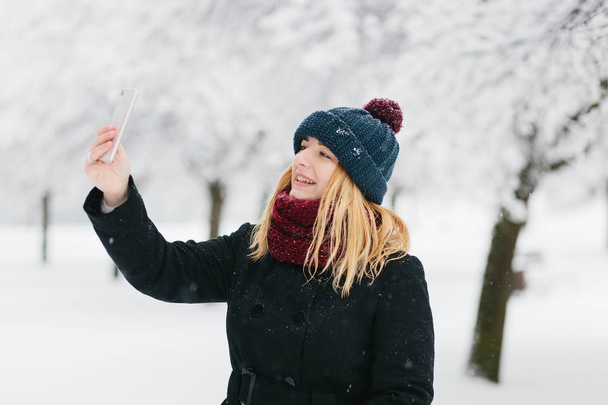 Jonge Kaukasische blond meisje in een zwarte jas en rode gebreide sjaal wandelingen in winter park met smartphone - Foto, afbeelding