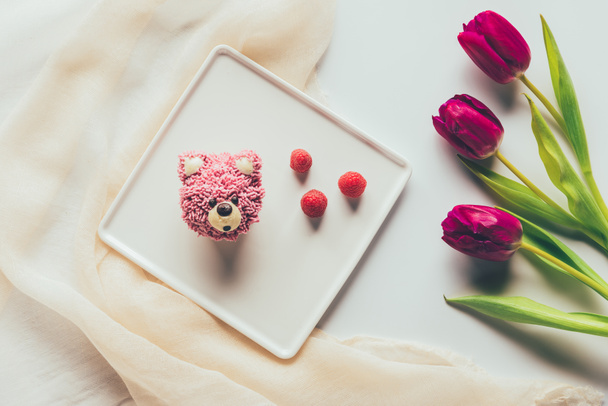 top view of sweet tasty muffin in shape of bear, fresh raspberries and tulip flowers   - Photo, Image