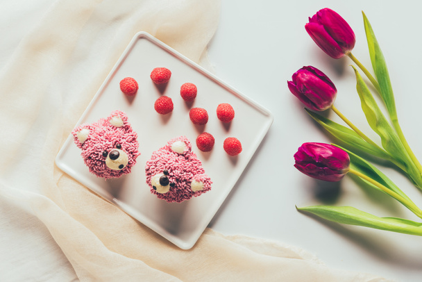top view of sweet tasty muffins in shape of bears, fresh raspberries and tulip flowers   - Фото, зображення
