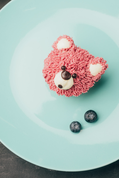 top view of sweet pink muffin in shape of bear and fresh blueberries on blue plate - Photo, Image
