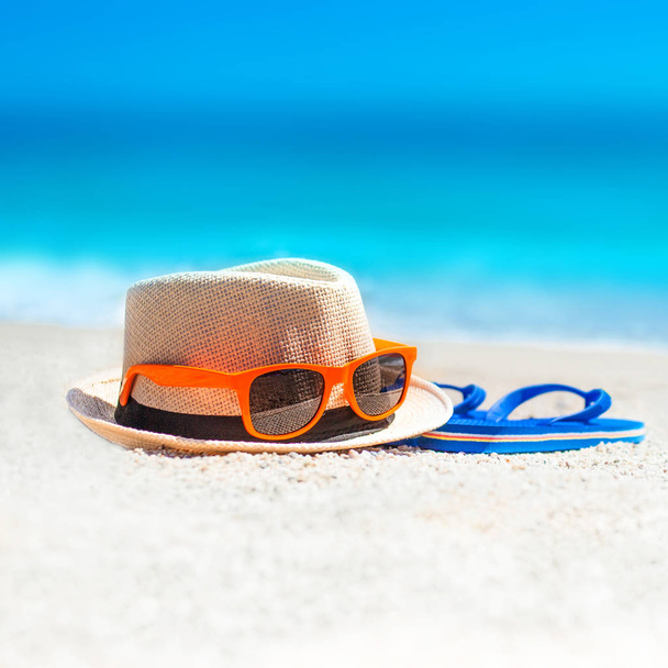 Chanclas, sombrero de playa, gafas de sol en la arena. Concepto vacaciones de verano. Día soleado en la playa tropical con horizonte azul del mar
 - Foto, Imagen