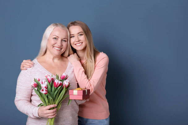 Young daughter with mother holding bouquet of flowers and gift box on color background - Foto, Imagem