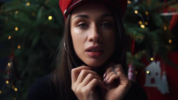 Charming model poses in red winter hat before a Christmas tree - Πλάνα, βίντεο