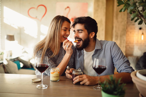 Hermosa pareja enamorada está sentada en la cafetería, bebiendo vino tinto y comiendo pastel. La joven está alimentando a su hombre. Mirándose suavemente el uno al otro
. - Foto, Imagen