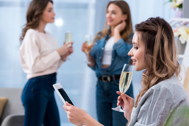 mujer joven sosteniendo una copa de champán y usando un teléfono inteligente mientras sus amigos hablan detrás
 - Foto, Imagen
