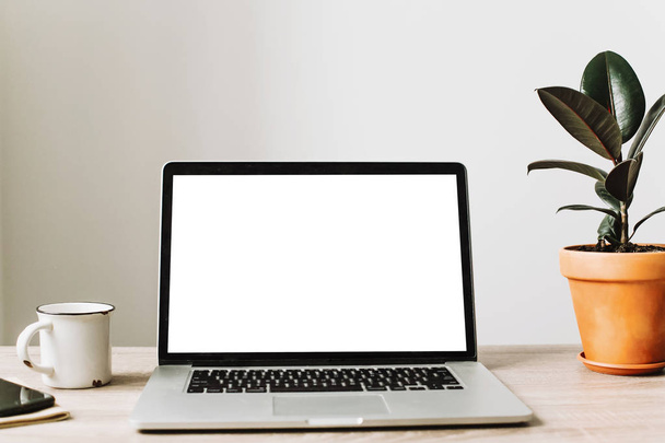 Laptop mit weißem leeren Bildschirm auf hölzernem Schreibtisch mit Telefon, Notizbuch, Kaffee und Pflanzen.  - Foto, Bild