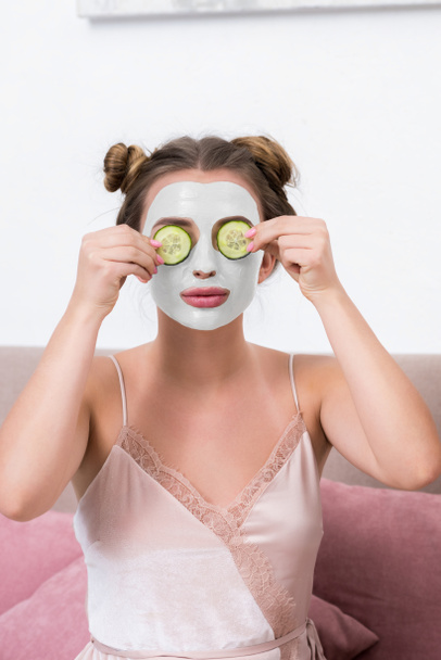 привлекательная молодая женщина в пижаме держит огуречные ломтики возле лица с маской
 - Фото, изображение