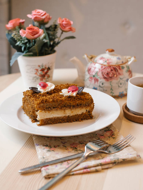 Torta al miele con crema tenera servita su piatto bianco con forchette, teiera, tazza di tè e fiori sullo sfondo
 - Foto, immagini