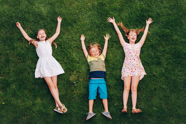 Niños felices divirtiéndose al aire libre. Niños jugando en el parque de verano. Niño y dos niñas tumbados sobre hierba fresca verde
 - Foto, imagen