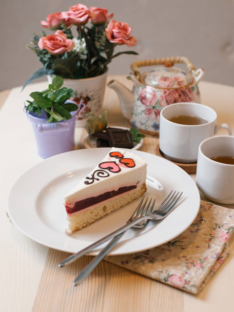 Biskuitteig mit Joghurtfüllung, Beerenmarmelade und dekorativem Zuckerguss, serviert auf weißem Teller mit Gabeln, Tassen Tee, Teekanne und Vasen - Foto, Bild