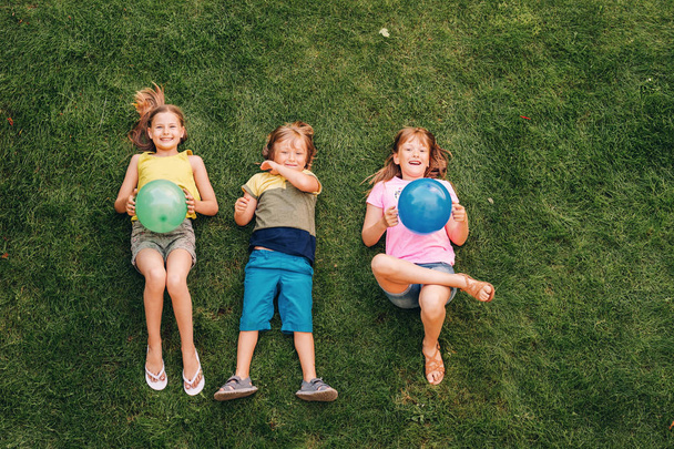 Des enfants heureux qui s'amusent dehors. Des enfants jouent dans un parc d'été. Petit garçon et deux filles couchés sur de l'herbe verte fraîche, tenant des ballons
 - Photo, image