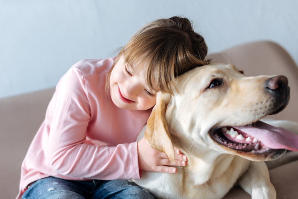 Happy child with down syndrome and Labrador retriever dog cuddling on sofa - Photo, Image