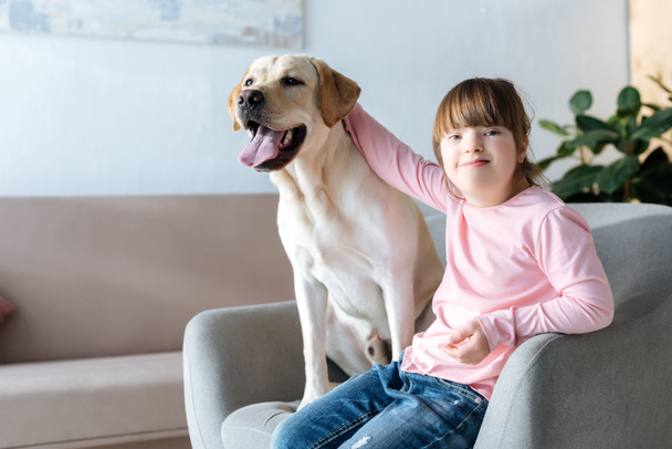 Kid with down syndrome stroking Labrador retriever dog sitting in chair - 写真・画像