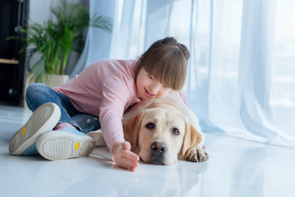 Child with down syndrome playing with Labrador retriever dog on the floor - Φωτογραφία, εικόνα