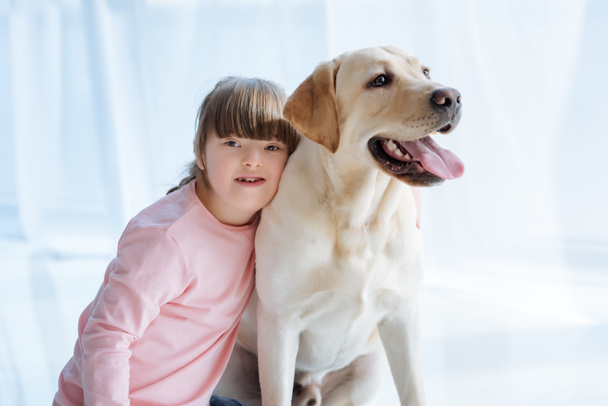Kid girl with down syndrome embracing Labrador retriever on light background - Photo, Image