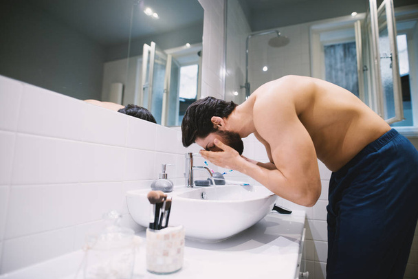 strong muscular shirtless man washing face in bathroom - Photo, Image
