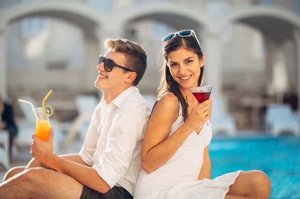 Two attractive people having cocktails by the pool in the summer vacation resort.Pool lounge bar party.Couple on exotic travel vacation enjoying refreshing drinks and relaxing day.Friends having fun - Фото, зображення