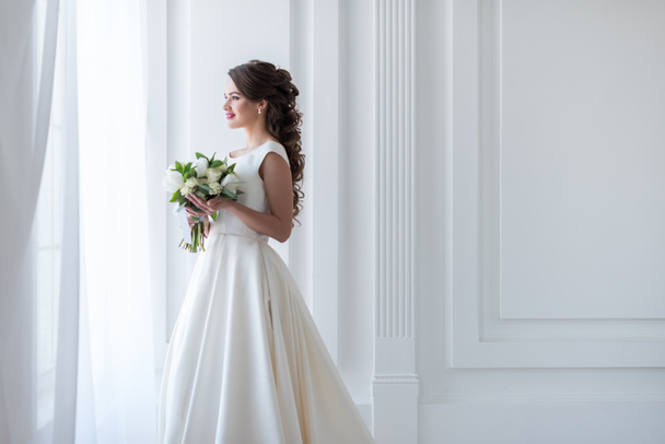 привлекательная счастливая невеста позирует в традиционном платье со свадебным букетом
 - Фото, изображение