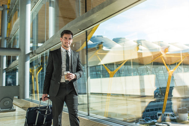 молодой бизнесмен прогуливается по терминалу аэропорта с багажом, улыбаясь кофе
  - Фото, изображение