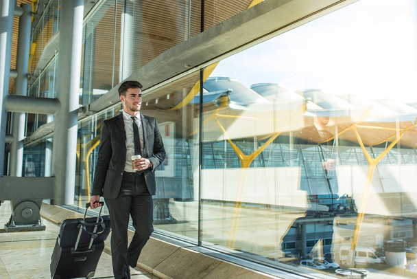 jonge zakenman wandelen in de luchthaventerminal met bagage glimlachend met een kopje koffie  - Foto, afbeelding