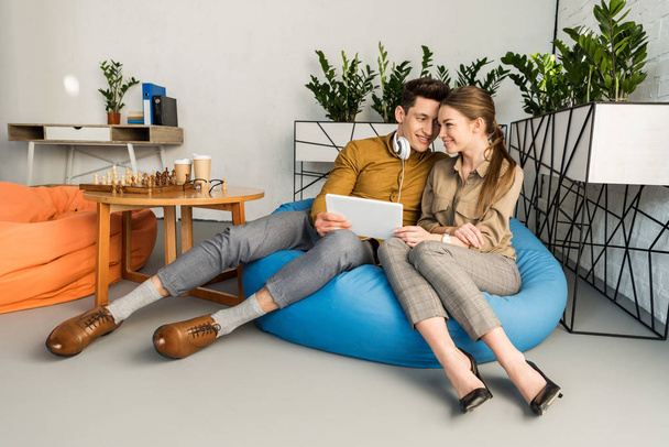 pareja joven abrazándose usando tableta juntos mientras están sentados en la bolsa de frijoles
 - Foto, Imagen